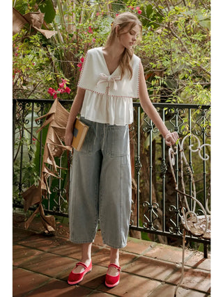 Woman standing on a balcony with plants and a book, wearing a white top and light blue pants.