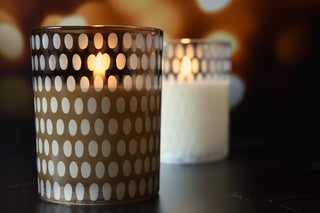A lit candle in a clear glass holder with a white oval dot pattern, placed on a dark surface with another similar candle blurred in the background against warm bokeh lights.