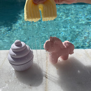 Yellow squirt toy spraying water onto purple and pink bath bombs by a pool.