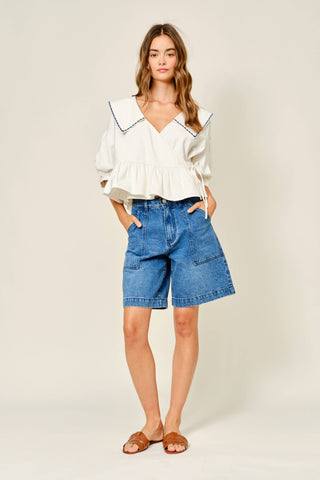 Woman wearing a white blouse with black trim and blue denim shorts on a beige background