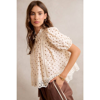 Woman wearing a white blouse with brown polka dots and brown pants against a neutral background