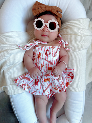 Baby in a red and white outfit with sunglasses and a bow headband on a white cushion.