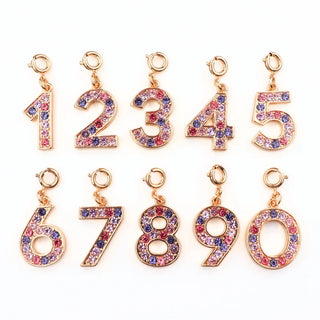 Ten gold-plated number charms from 1 to 0, each embellished with pink, purple, and blue rhinestones, arranged in two rows on a white background.