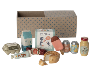 A miniature toy food and grocery play set arranged in front of a cardboard storage box. The set includes toy eggs, milk carton, ice cream carton, popsicle, canned drinks, honey bottle, mustard bottle, sausage, bread, and a tube of mayonnaise on a white background.