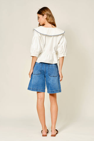 Woman wearing a white blouse with a boat neckline and blue denim shorts on a plain background