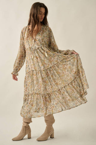 A woman modeling a long-sleeve, tiered maxi dress with a beige and brown floral print, paired with beige heeled boots, against a solid light beige background.