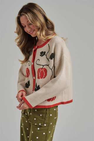 A woman wearing a cream knit cardigan with red trim and black-and-red pumpkin motifs, paired with olive green pants with white polka dots, against a solid gray studio background.