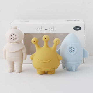 Three colorful toy figures in front of an 'ali+oli' product box on a white background