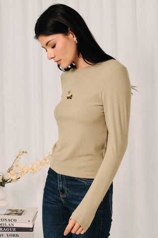 A woman wearing a beige long-sleeve crewneck sweater with a small embroidered bee detail on the left chest, paired with dark blue jeans, standing against a white curtain background.
