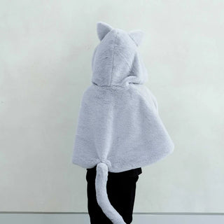 A person shown from the back wearing a light gray plush hooded cape with cat ears and a long tail, paired with black pants, against a plain off-white wall.
