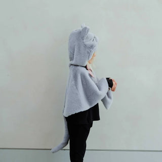 A child wearing a light gray plush cat costume cape with a tail and cat ears on the hood, standing against a plain light gray wall.