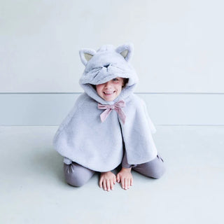 A smiling child kneeling on a light floor wearing a plush light gray hooded cape with cat ears and a pink bow, paired with gray leggings.