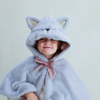 A child wearing a plush light blue cat-themed hooded cape with gold inner ears and a dusty pink bow at the neck, photographed against a soft pale green background.
