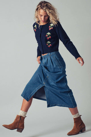 Woman wearing a navy sweater with floral embroidery and a blue denim skirt on a plain background