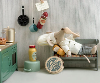 Toy rabbit lying on a small bed in a miniature kitchen setting with various kitchen items around.