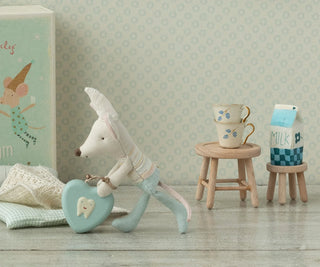 Stuffed toy mouse with a heart-shaped object in a room with a patterned wall and small wooden stool with cups and milk carton.