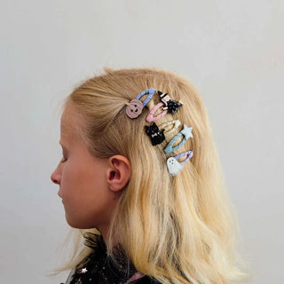 A side-profile view of a young girl with blonde hair wearing a set of colorful, whimsical hair clips, including a button, a ghost, a star, and a small cat, against a plain light gray background.