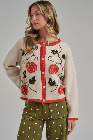A woman models a cream-colored knit cardigan featuring red trim and a black vine pattern with red pumpkins, paired with olive green polka dot pants against a solid gray studio background.