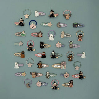 A collection of small Halloween-themed hair clips arranged in rows on a muted blue background. The clips feature cute designs including witches, ghosts, pumpkins, owls, skeletons, and broomsticks in soft pastel and earthy tones.