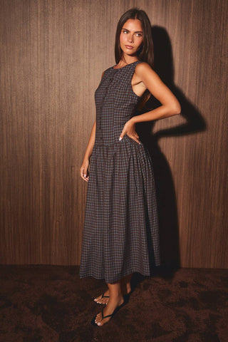 A woman modeling a sleeveless midi dress with a dark gray and black gingham pattern, standing against a dark wood-paneled wall.