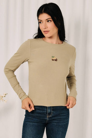 A woman modeling a beige long-sleeve crewneck top with a small embroidered cherry design on the left chest, paired with dark blue jeans against a white curtain background.