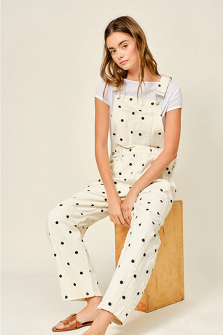 Woman wearing a white polka dot jumpsuit sitting on a wooden block against a beige background