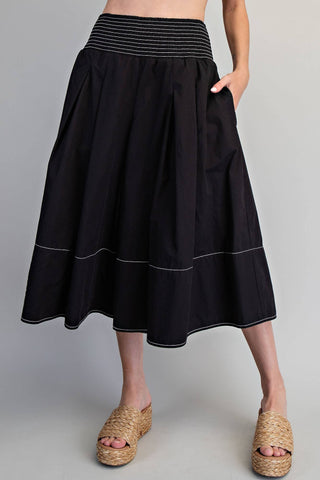 A black midi skirt with a striped elastic waistband and white contrast stitching, shown on a model wearing woven platform sandals against a neutral gray background.