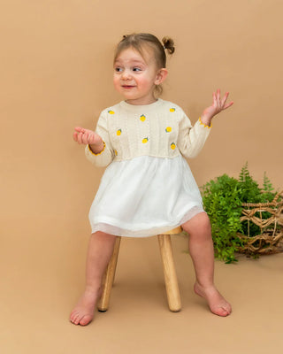 Young girl in a white dress with yellow accents sitting on a wooden stool against a beige background.