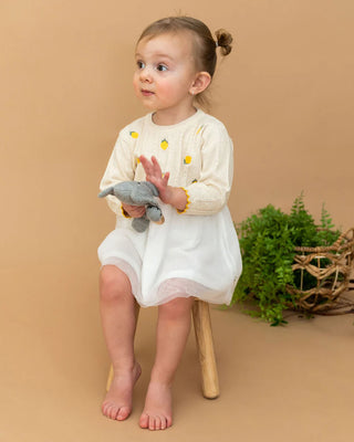 Child in a white dress with yellow flowers holding a gray toy on a brown background