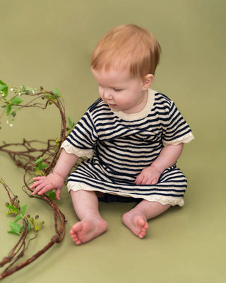 Baby in a striped dress sitting on a green background with branches.