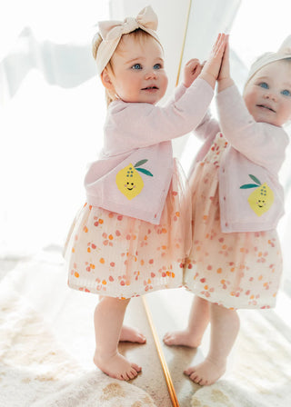 Baby in a pink outfit with lemon design standing in front of a mirror.