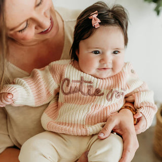 Woman holding a baby wearing a pink and white checkered sweater with 'Cutie pie' on it.