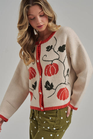 A woman wearing a cream-colored knit cardigan with red trim and a black and red pumpkin vine pattern, paired with olive green pants with white polka dots, against a solid gray studio background.