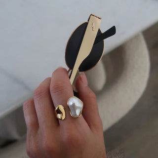 Hand holding a pair of sunglasses with gold frame and black lenses, wearing a pearl ring.