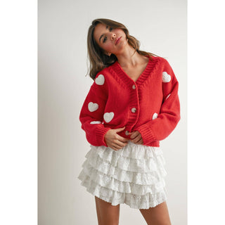 Woman wearing a red cardigan with white heart patterns and a white lace skirt on a plain background