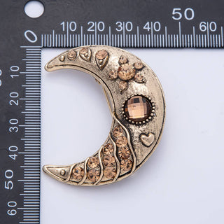Decorative brass moon-shaped pendant with gemstones on a ruler for scale.