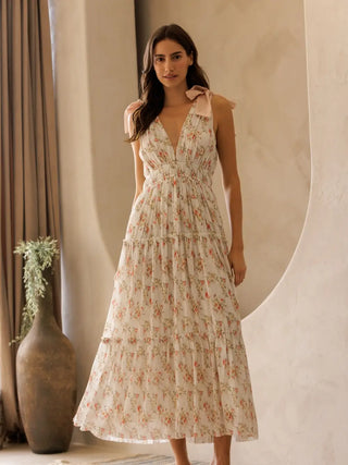 Woman wearing a floral dress standing in a softly lit room with a vase and plant in the background.