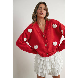 Woman wearing a red cardigan with white heart patterns and a white skirt.