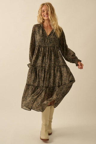 A woman modeling a long-sleeve, tiered midi dress with an abstract dark brown and olive pattern, paired with beige ankle boots, against a solid beige studio background.