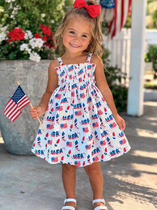 Child wearing a dress with American flag patterns, holding a small American flag, outdoors.