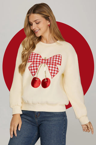 Woman wearing a cream sweater with a red and white checkered bow and cherry design against a gray background.