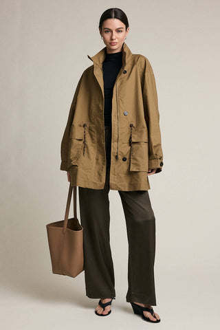 Woman wearing a beige coat and dark pants, holding a brown bag on a plain background