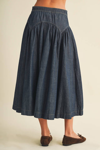 A dark blue denim midi skirt with a gathered waist and pleated details, shown on a model from the back wearing brown suede flats against a light beige background.