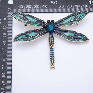 Dragonfly-shaped brooch with teal and gold details on a white background with a ruler for scale.