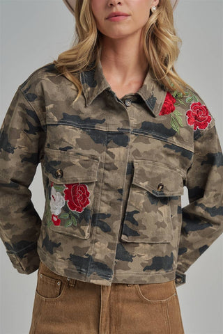A woman wearing a cropped camouflage jacket with red rose embroidery on the chest and shoulders, paired with brown pants, photographed against a neutral gray studio background.