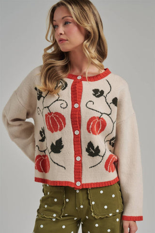 A woman wearing a cream knit cardigan with red trim and a red, black, and green pumpkin vine pattern, paired with olive green polka-dot pants, against a solid gray studio background.