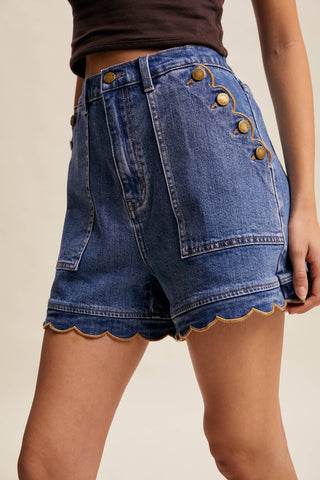 Blue denim shorts with scalloped hem and gold buttons on a beige background