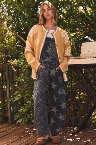 A woman stands outdoors on a wooden deck wearing a mustard yellow knit cardigan with white leaf patterns, blue denim overall covered in gray star prints, and tan slip-on shoes, with dense green foliage behind her.
