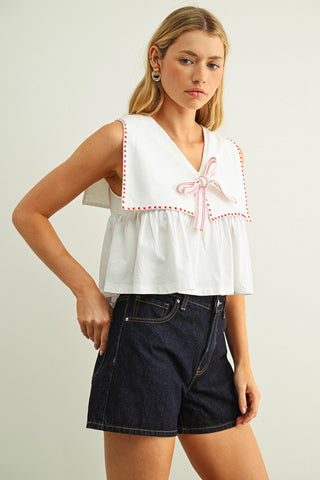 Woman wearing a white sleeveless top with pink trim and dark denim shorts on a plain background