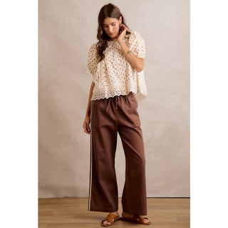 Woman wearing a white blouse with lace details and brown pants against a beige wall.